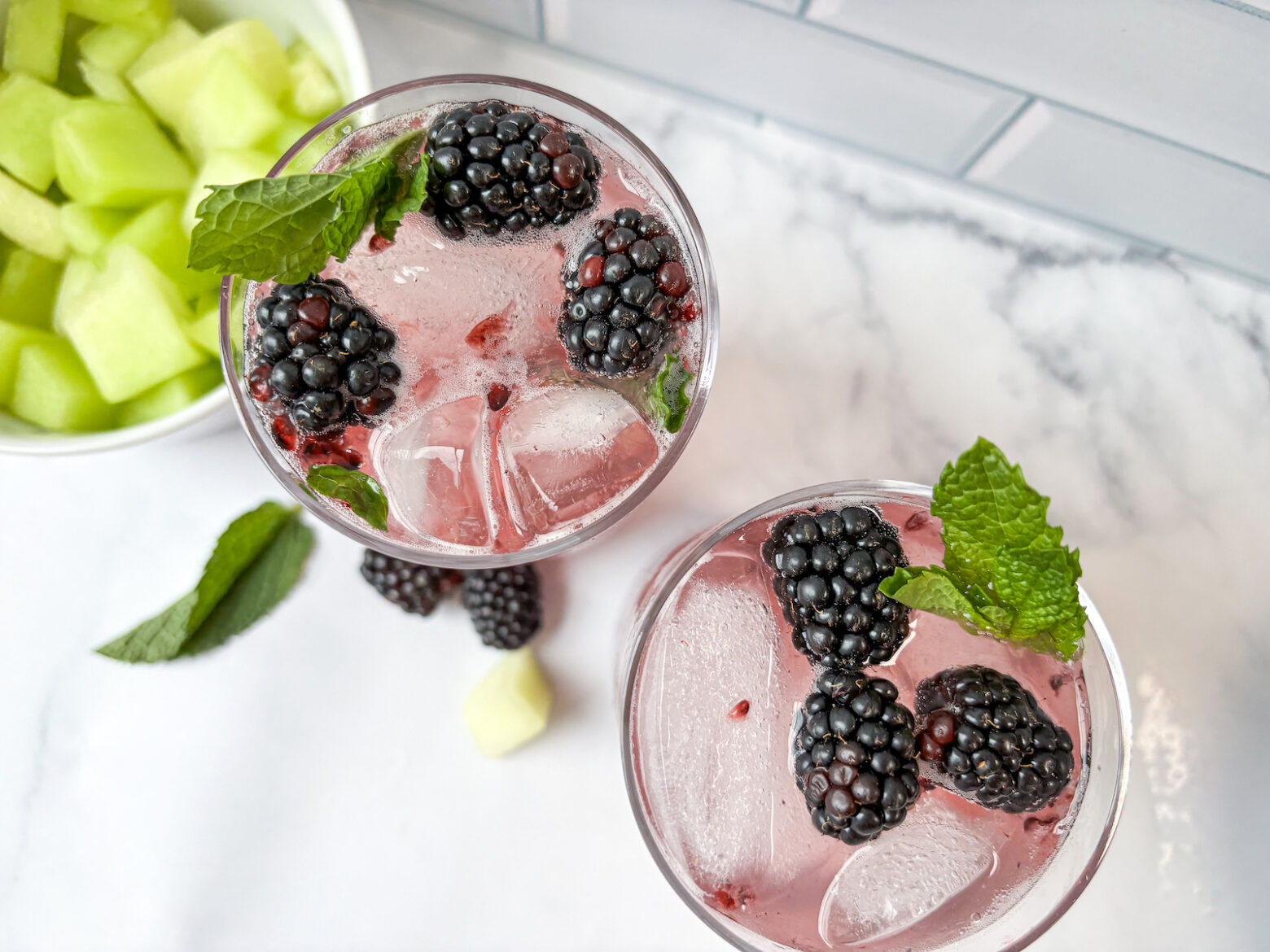 Birds eye view of two honeydew watermelon mocktails sitting diagonal to each other with garnishes and melon in the background