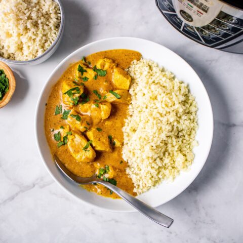 From above, a bowl of half butter chicken garnished with chopped parsley with a silver spoon and half cauliflower rice.