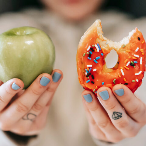 Close-up of a woman holding up a green apple and an orange-frosted donut with a bite taken out of it