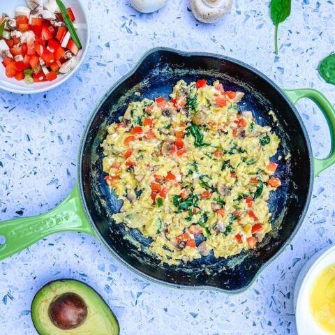 From above, an iron skillet with scrambled eggs and vegetables, surrounded by a bowl of chopped uncooked vegetables, two raw mushrooms, spinach leaves, half an avocado and a cracked raw egg in a bowl.