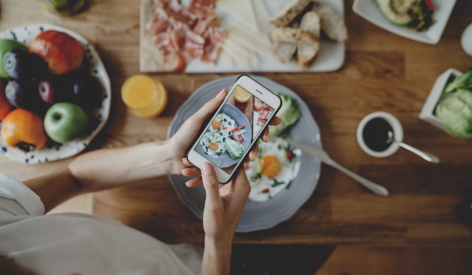 From above, a woman photographs her meal on a table surrounded by food.