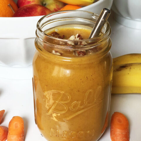 A mason jar full of pumpkin pie smoothie with a silver metal straw, garnished with chopped pecans and surrounded by a large white bowl of fruit, two bananas and some carrots.