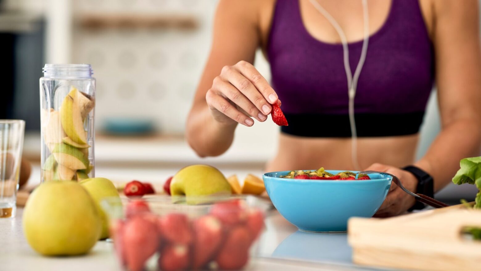 A woman in work-out attire makes herself a bowl of fruit.