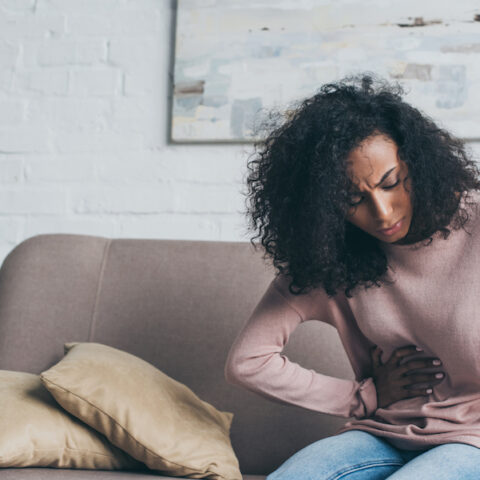 A woman in pain sitting on a couch with her hand on her stomach.